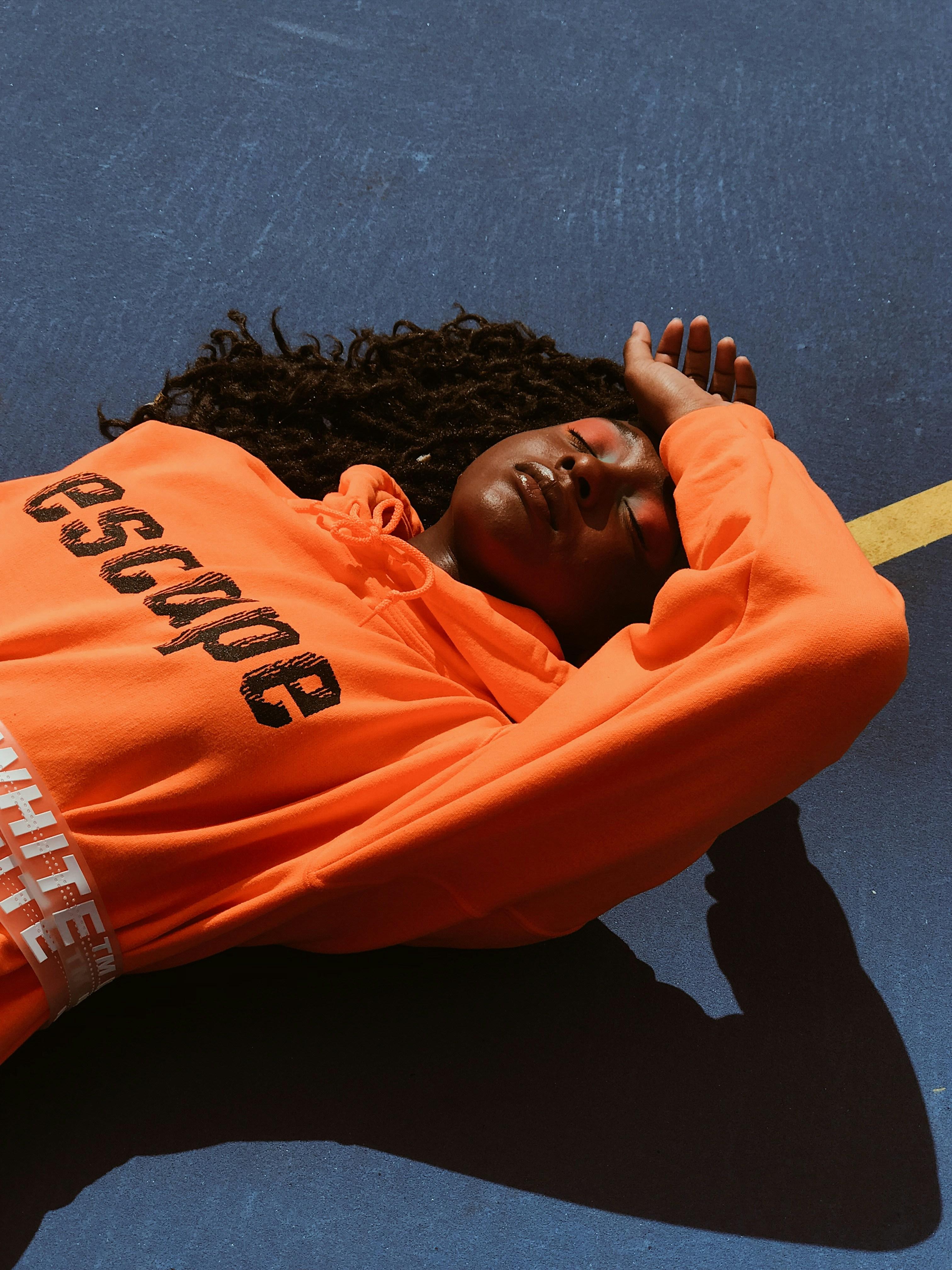 Image of a woman laying down in the sun with her eyes closed, wearing an orange hoodie and orange eyeshadow.